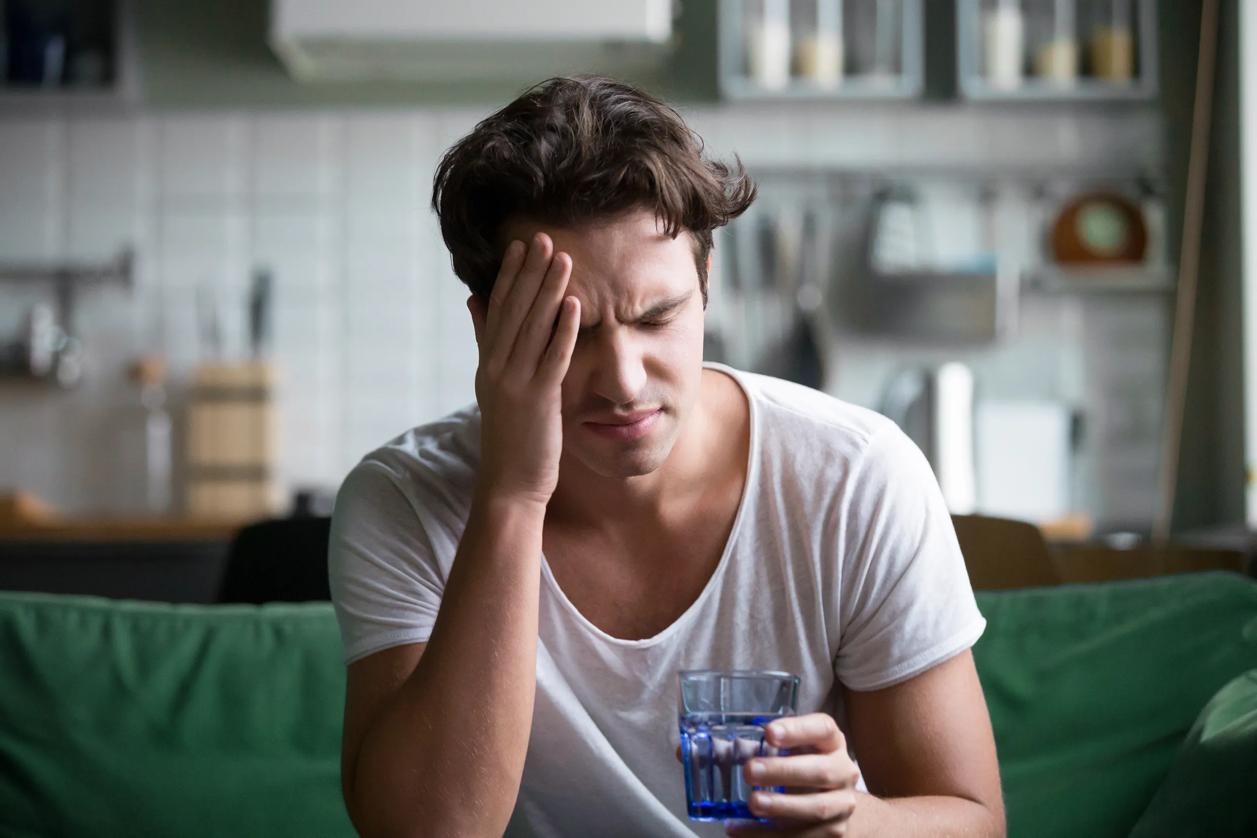 Young man in a white tshirt sitting on a green couch in an apartment holding his head with a glass of water