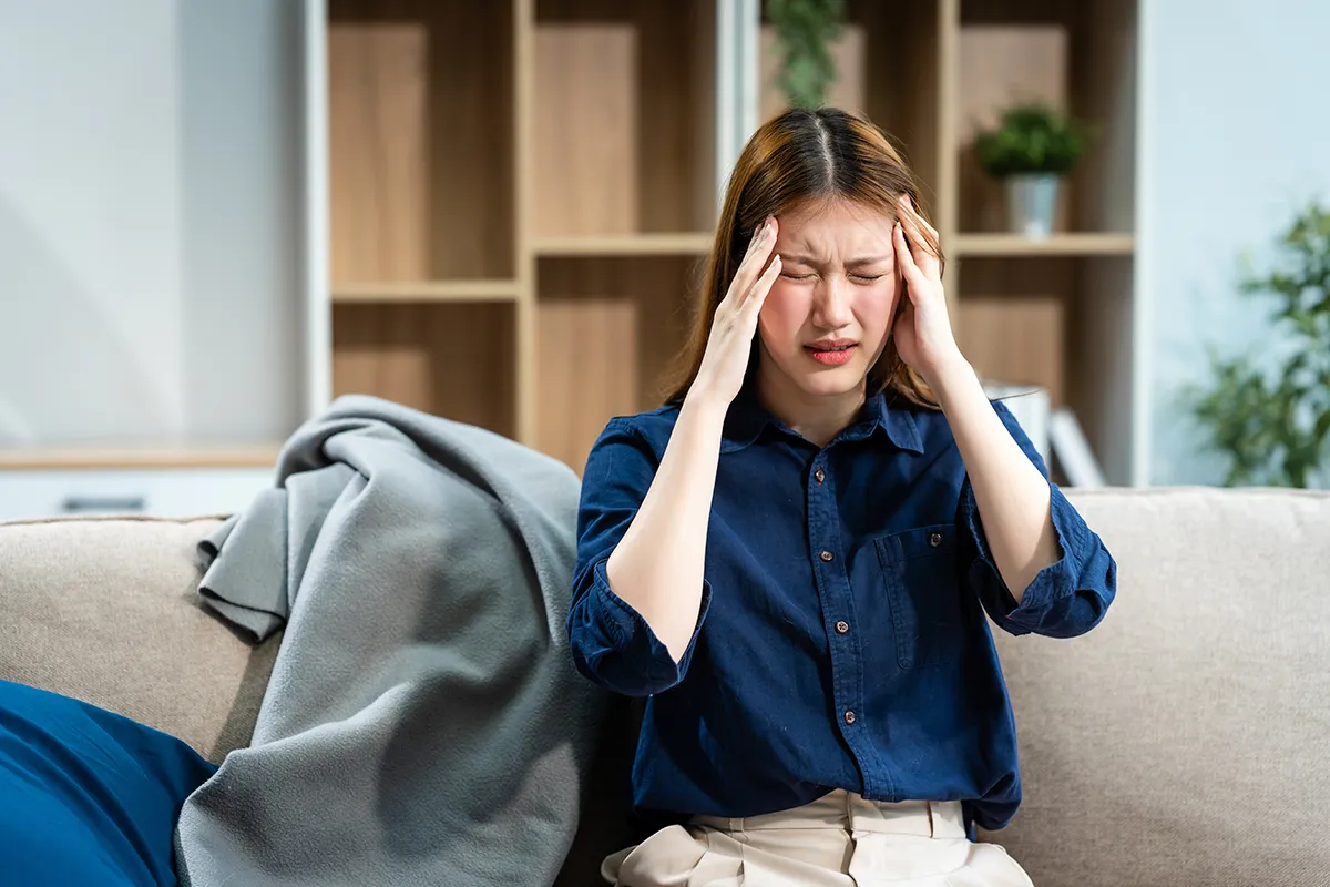 Young Asian woman sitting on a lounge holding her head - headache, migraine pain